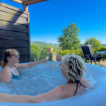 Genießen Sie ein Glas Sekt im sprudelnden Whirlpool mit Ausblick auf grüne Hügel und blauen Himmel.