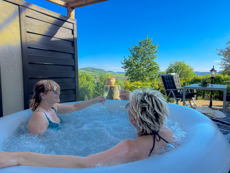 Genießen Sie ein Glas Sekt im sprudelnden Whirlpool mit Ausblick auf grüne Hügel und blauen Himmel.