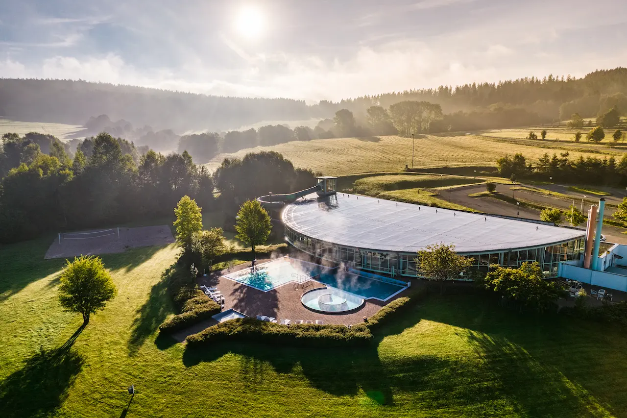 Luftaufnahme des Inselbergbads Brotterode mit Außenbecken, umliegenden Feldern und Wald im Morgenlicht.