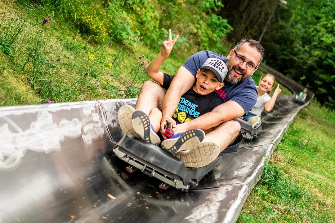 Vater und Kind rodeln lachend die Sommerrodelbahn im Inselsberg Funpark hinab, weitere Besucher folgen dahinter.