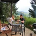 Zwei Erwachsene sitzen an einem Tisch auf einer überdachten Terrasse. Sie essen gemeinsam. Rechts steht ein Kugelgrill. Im Hintergrund sieht man Bäume und eine hügelige Landschaft.