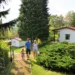Eine dreiköpfige Familie – Vater, Mutter und ein kleines Kind – geht an einem sonnigen Tag lächelnd einen gepflasterten Gartenweg in einer hügeligen Ferienhausanlage hinauf. Im Hintergrund sind zwei weiße Bungalows mit roten Dächern zwischen dichten Nadelbäumen und grünen Sträuchern zu sehen.