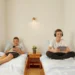 Ein helles Schlafzimmer mit zwei separaten Einzelbetten aus Holz. Auf dem linken Bett liegt ein Junge entspannt auf dem Rücken und schaut auf sein Smartphone. Auf dem rechten Bett sitzt ein anderer Junge im Schneidersitz, trägt Kopfhörer und hält ein Tablet in den Händen. Zwischen den Betten steht ein kleiner Nachttisch aus Holz mit einer Pflanze. Ein großes Fenster mit Jalousien auf der linken Seite sorgt für viel natürliches Licht.