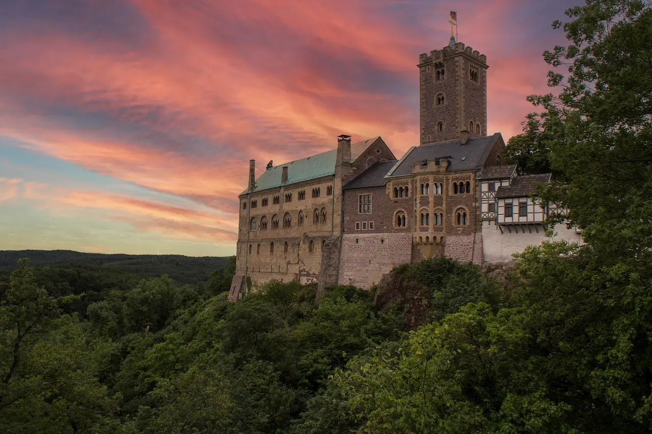 Die Wartburg in Eisenach, UNESCO-Weltkulturerbe in Thüringen, vor dramatischem Sonnenuntergang mit bewaldeter Umgebung.
