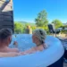 Ein Paar genießt ein Glas Sekt im sprudelnden Whirlpool mit Ausblick auf grüne Hügel und blauen Himmel.