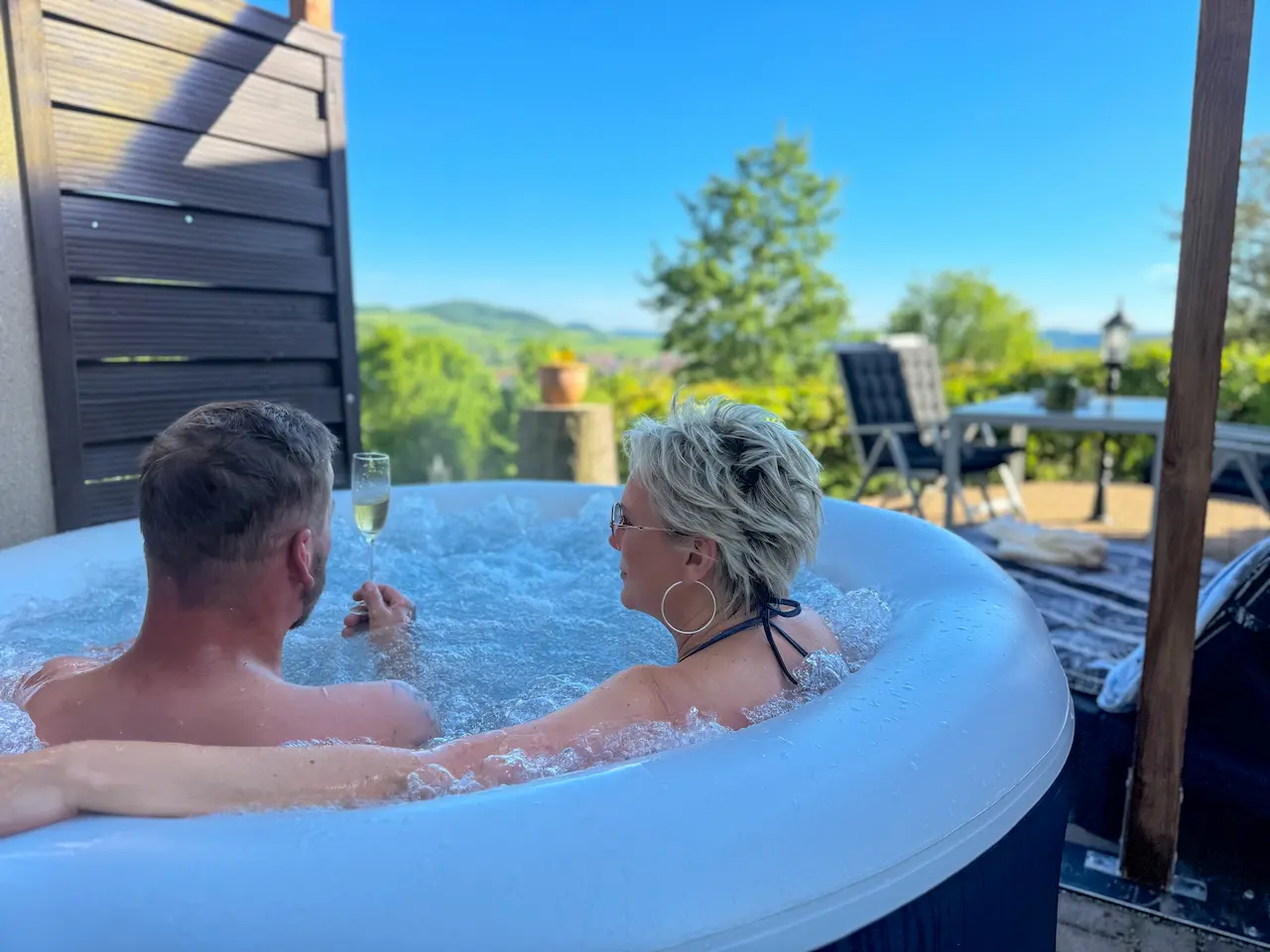 Ein Paar genießt ein Glas Sekt im sprudelnden Whirlpool mit Ausblick auf grüne Hügel und blauen Himmel.